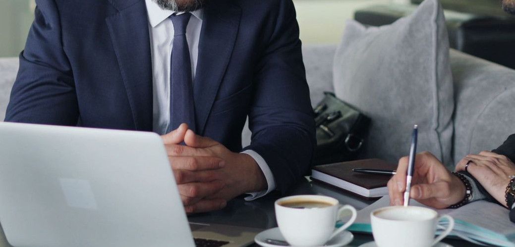Two businessmen in suits discussing work at a cafe with laptops and coffee.